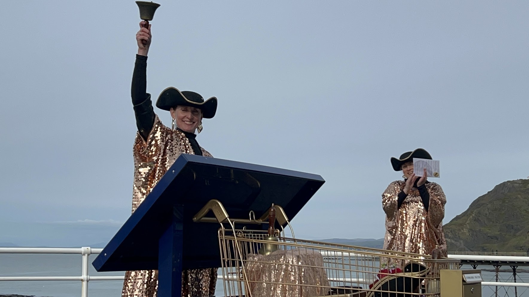 Dr Louise Ritchie mewn gwisg aur yn galw llythyr o gariad, gyda chloch yn ei llaw, ar y prom yn Aberystwyth fel rhan o brosiect Er Mwyn Dyn